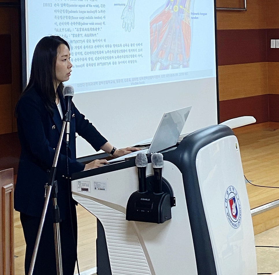 한 여성이 강단에서 노트북과 마이크 앞에서 발표하고 있으며, 뒤 스크린에는 해부학 그림과 글이 보인다.
