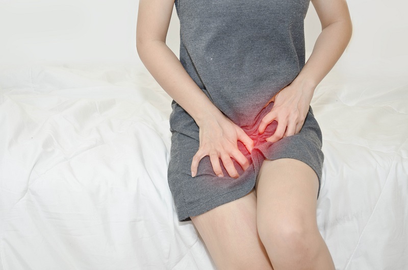 흰 침대에 앉아 회색 옷을 입은 여성이 사타구니 부위를 양손으로 긁고 있으며, 해당 부위에 붉은색 강조 표시가 있다 — 서울 바로한의원 초음파 약침·도침