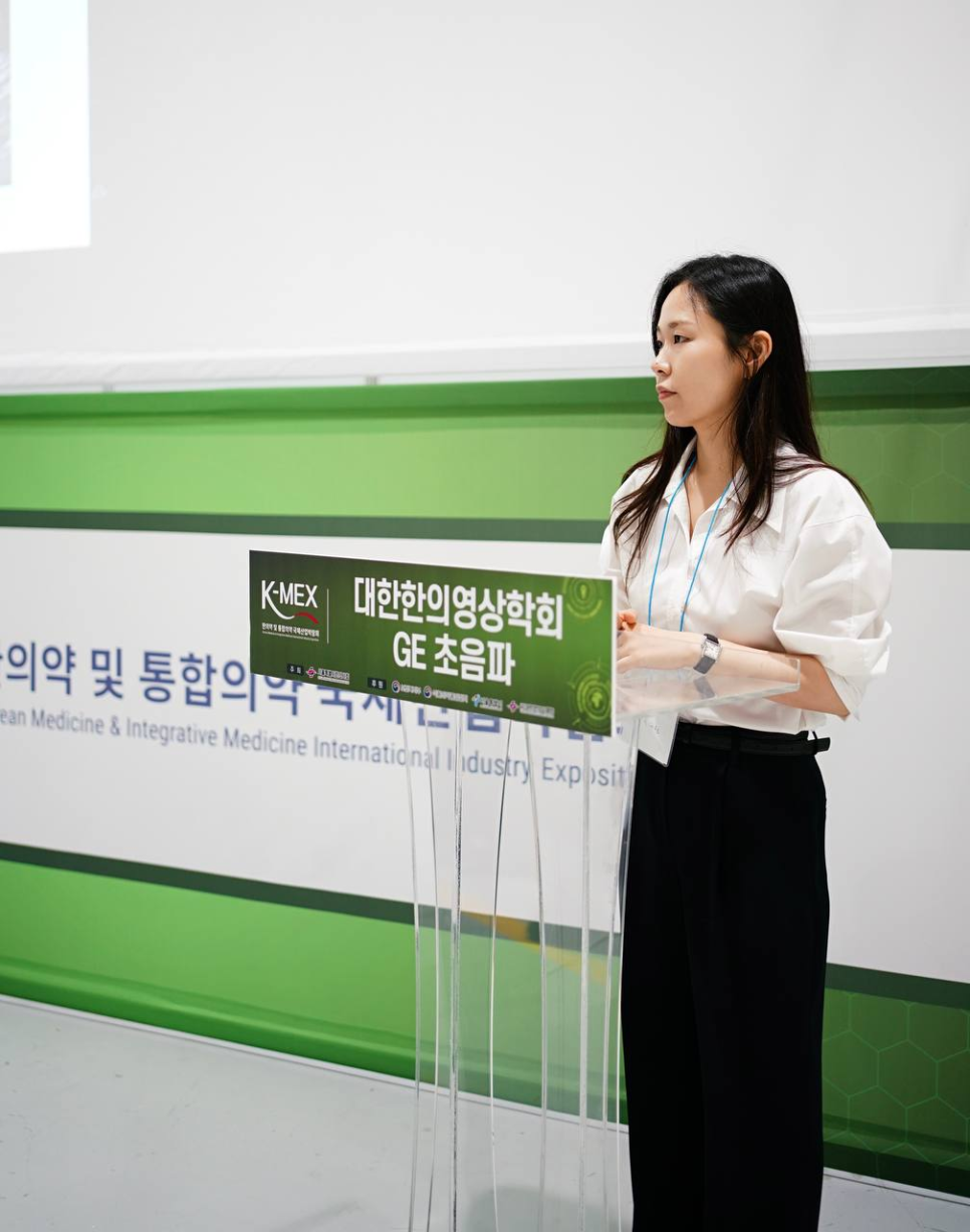 흰 셔츠를 입은 여성이 '대한한의영상의학회 GE 초음파'라고 적힌 투명 연단 뒤에 서 있다.