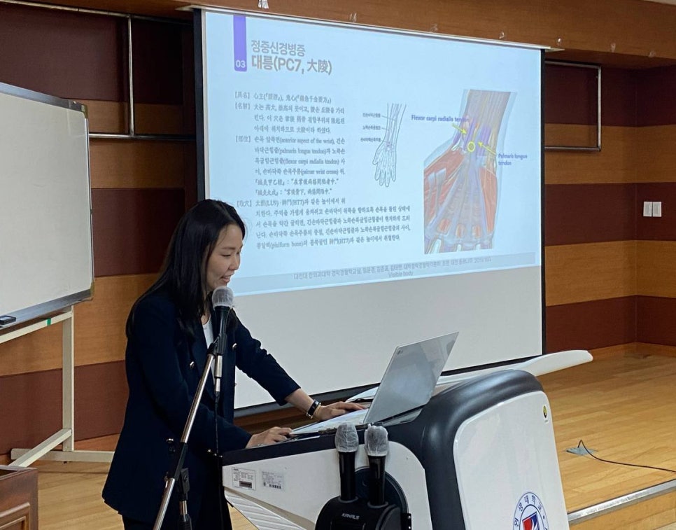 한 여성이 강단에서 노트북과 마이크가 있는 연단에 서서 발표하고 있으며, 뒤편 스크린에는 손목 해부학 다이어그램과 정중신경병증 관련 한글 텍스트가 보인다.