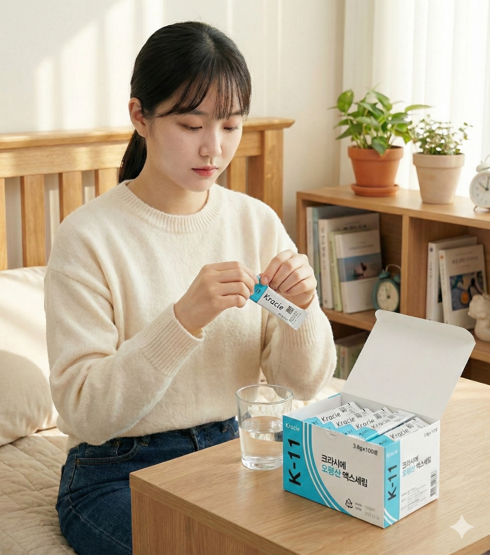 한 여성이 테이블에 앉아 크라시에 오령산 스틱 포를 뜯고 있으며, 옆에는 물 한 잔과 제품 상자가 놓여 있다.
