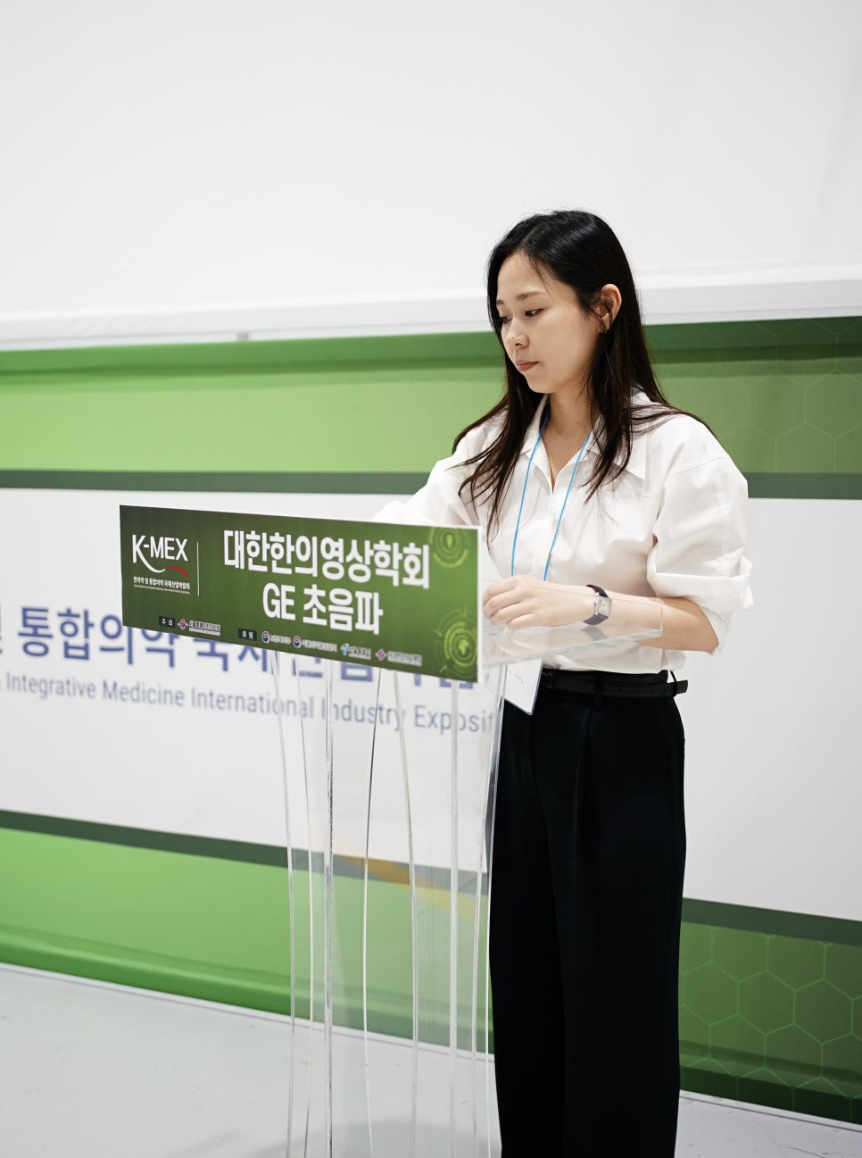 여성이 투명 연단에 서서 아래를 보고 있으며, 연단에는 '대한한의영상학회 GE 초음파'라고 적힌 표지가 놓여 있다.