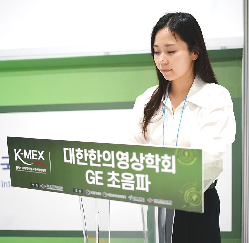흰 셔츠를 입은 여성이 K-MEX 대한한의영상의학회 GE 초음파라고 적힌 연단 뒤에 서 있다.