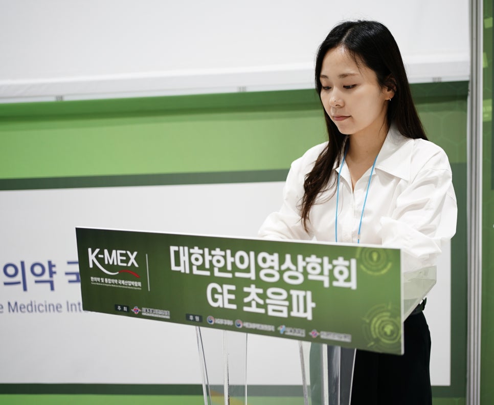 K-MEX와 대한한의영상학회 GE 초음파라고 적힌 연단에 서서 아래를 보고 있는 여성