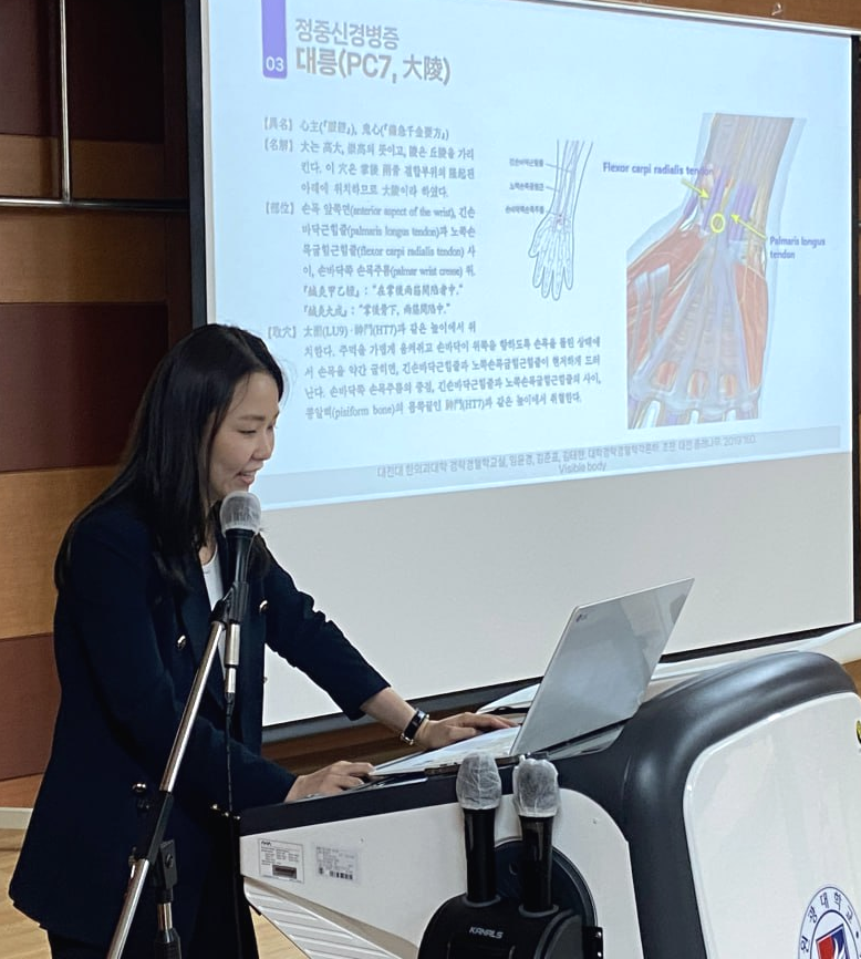한 여성이 강단에서 발표하며, 뒤쪽 스크린에 손목 해부학 다이어그램과 한의학 관련 내용이 보인다.