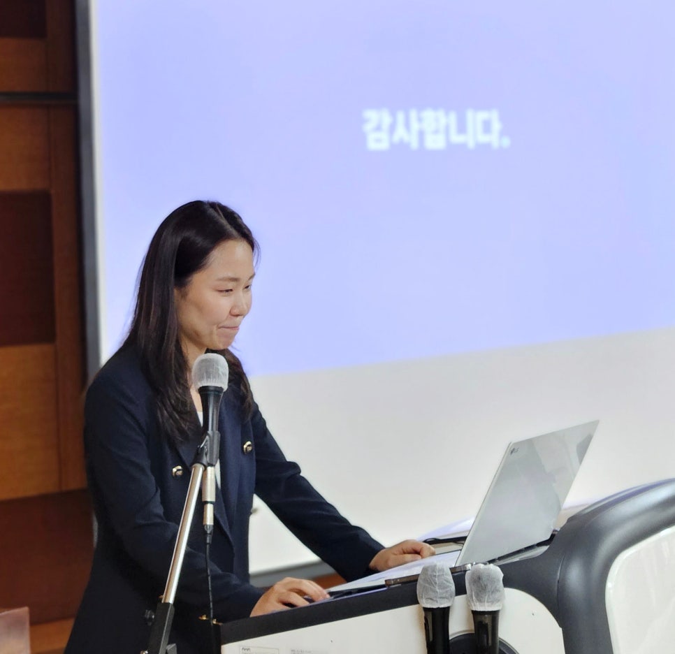 강단에서 마이크와 노트북을 앞에 두고 발표하는 여성의 모습.