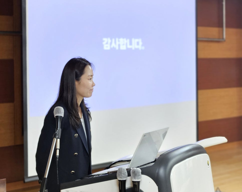 마이크가 놓인 강단에 서서 옆을 바라보는 여성. 뒤편 화면에는 '감사합니다'라는 문구가 보인다.