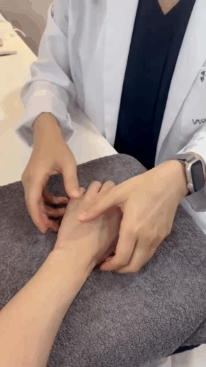 흰색 가운을 입은 사람이 다른 사람의 손목과 손을 만져 진찰하는 모습.