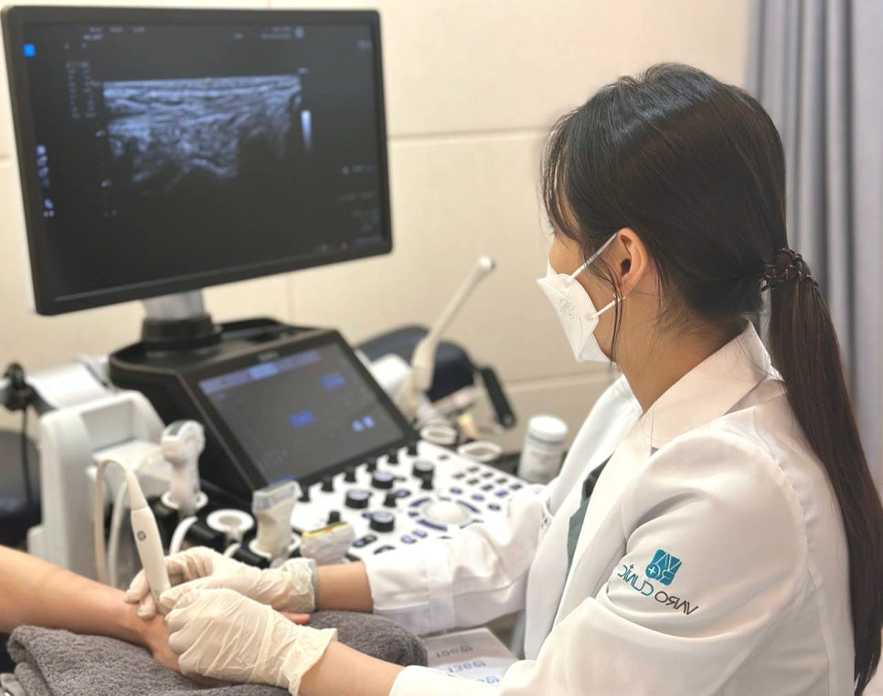 마스크를 쓴 의료진이 환자의 손목에 초음파 기기를 대고 검사하며, 모니터에 초음파 영상이 보인다.